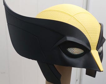 May include: A black and yellow plastic costume mask with a textured surface. The mask has a large, black, molded eye area with a white mesh covering the eyes. The mask is designed to resemble the character Wolverine.