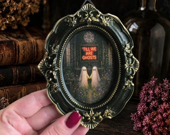 May include: An oval wall hanging featuring a black frame with gold-toned ornate detailing. The central artwork depicts two white ghost figures holding hands in a dark room with the orange neon text 'TILL WE ARE GHOSTS' displayed above them.