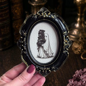 Puede incluir: Un marco de fotos ovalado de color negro con detalles ornamentados en tono dorado, que contiene una pequeña ilustración de una figura de fantasma blanca sosteniendo una pila alta de libros oscuros. El marco presenta un borde interior con cuentas y un borde exterior decorativo y texturizado.