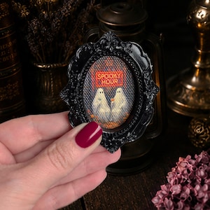 May include: A small oval black ornate frame containing a cross-stitch embroidery of two white ghosts. The ghosts are positioned side by side, and the text "SPOOKY HOUR" is stitched in orange letters above them on a dark textured background.