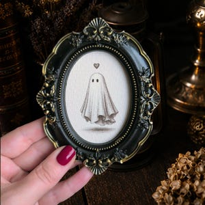 Può includere: Una cornice ovale con un bordo decorato nero e oro contenente un piccolo disegno di un fantasma con un minuscolo cuore sopra la testa. La cornice presenta intricati dettagli in rilievo e un bordo interno perlato che circonda l'illustrazione.