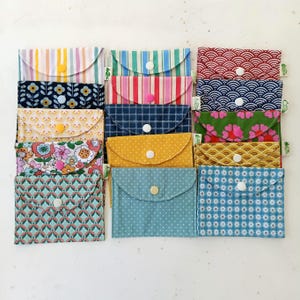 Peut inclure: Une collection de quinze petites pochettes en tissu disposées en grille, chacune présentant un motif unique incluant des rayures, des fleurs, des pois et des formes géométriques. Chaque pochette possède un rabat fermé par un bouton-pression.