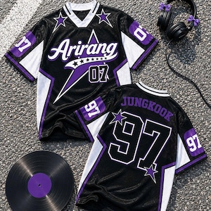 May include: Two black, white, and purple mesh jerseys featuring the text Arirang and 07 on the front, and JUNGKOOK and 97 on the back. The jerseys include star graphics, white V-neck collars, and white side panels. A vinyl record and black headphones are nearby.