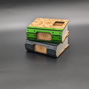 May include: Two wooden boxes stacked on top of each other. The top box is decorated with a swirling pattern and has a green stripe with black squares. The bottom box is gray with a black stripe. Both boxes have a circular design on the front.
