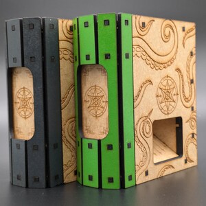 May include: Two dice towers made of wood with a black and green painted finish. The towers have a design of tentacles and a pentagram on the front. The towers are designed to hold dice for tabletop games.