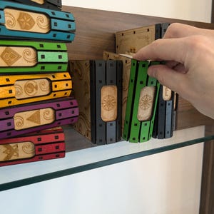May include: A stack of colorful books with decorative designs on the spines. The books are arranged on a shelf with a glass top. A hand is reaching for the books.