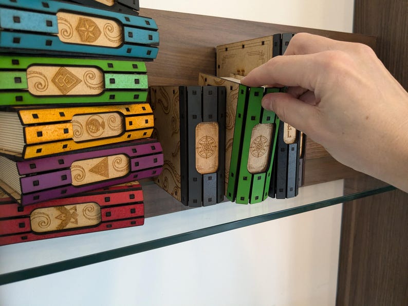 May include: A stack of colorful books with decorative designs on the spines. The books are arranged on a shelf with a glass top. A hand is reaching for the books.
