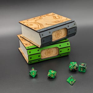 May include: Two wooden book-shaped boxes stacked on top of each other. The top box is brown with a black metal trim and the bottom box is green with a black metal trim. Both boxes have a circular design on the front. There are five green and gold dice on a black surface.