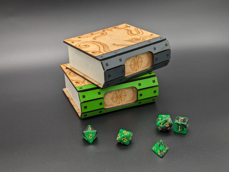 May include: Two wooden book-shaped boxes stacked on top of each other. The top box is brown with a black metal trim and the bottom box is green with a black metal trim. Both boxes have a circular design on the front. There are five green and gold dice on a black surface.