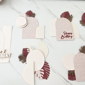 May include: Arrangement of layered paper cutouts in cream, burgundy, and brown, with floral patterns and tropical leaf accents. Some pieces have the words "Happy Birthday" in burgundy. The pieces are arranged on a white marble surface.