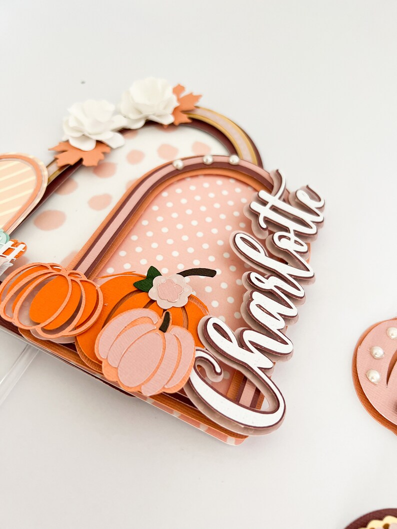 K&ouml;nnte beinhalten: Ein mehrschichtiger Tortenaufsatz mit Herbstthema. Der Aufsatz zeigt einen Rahmen mit gepunktetem Hintergrund, K&uuml;rbissen und dem Namen "Charlotte" in Schreibschrift. Wei&szlig;e Blumen und Perlen f&uuml;gen Details hinzu.