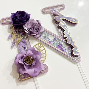 Può includere: Decorazione per torta a forma di lettera "N", con dettagli viola e oro. Il design include fiori di carta, foglie dorate e glitter. La lettera ha una sezione trasparente riempita di paillettes e piccole perle viola.