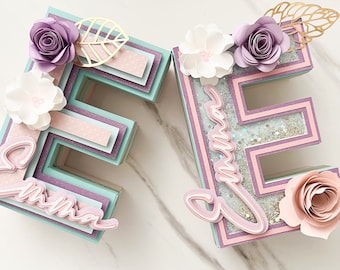 May include: Two decorative letter "E" boxes in pastel colors. The boxes are layered with light blue, lavender, and pink paper. Each letter is adorned with paper flowers, gold leaf accents, and the name "Emma" in cursive.