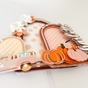 K&ouml;nnte beinhalten: Ein mehrschichtiges Papierkunstwerk mit einem rosa Pickup-Truck, K&uuml;rbissen und floralen Akzenten. Das Design umfasst bogenf&ouml;rmige Rahmen mit gepunkteten und gestreiften Mustern sowie das Wort "Hello" in Schreibschrift.