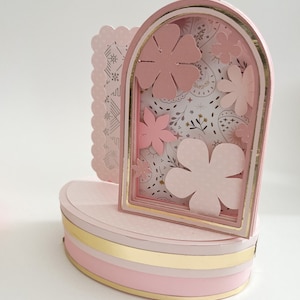 Può includere: Un'opera d'arte in carta a strati in rosa tenue e oro. Il design presenta un arco centrale con fiori di carta, su uno sfondo a motivi. La base è di forma arrotondata con strisce dorate e rosa e un bordo smerlato a sinistra.