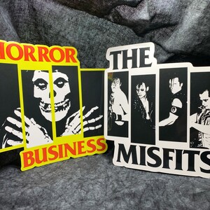 May include: Two black and white stickers with the band The Misfits logo. The first sticker says "Horror Business" with a skull in a prison cell. The second sticker says "The Misfits" with four band members.