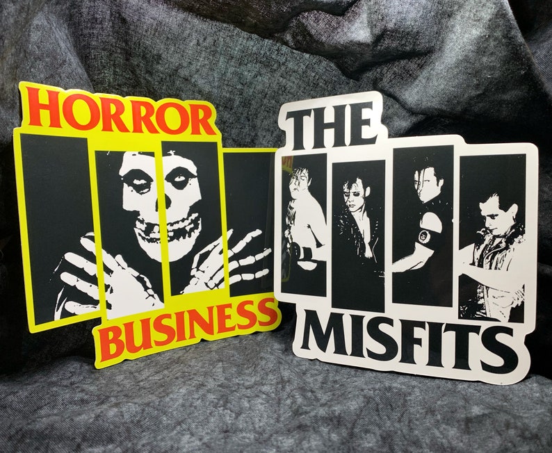 May include: Two black and white stickers with the band The Misfits logo. The first sticker says "Horror Business" with a skull in a prison cell. The second sticker says "The Misfits" with four band members.