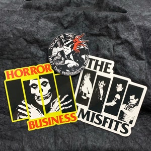 May include: Three black and white stickers with band logos. The top sticker is round and features a man with a microphone and the text "Texas is the reason that the presidents are dead". The middle sticker features a skull with the text "Horror Business". The bottom sticker features a band of four people with the text "The Misfits".