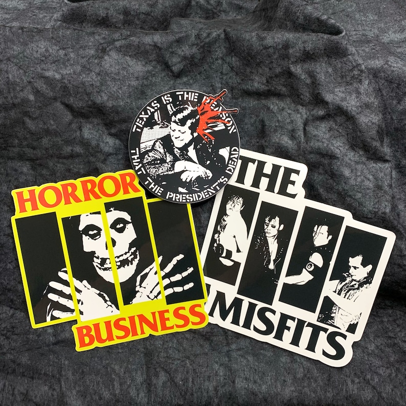 May include: Three black and white stickers with band logos. The top sticker is round and features a man with a microphone and the text "Texas is the reason that the presidents are dead". The middle sticker features a skull with the text "Horror Business". The bottom sticker features a band of four people with the text "The Misfits".