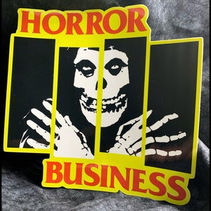 May include: A yellow and black sticker with the words "HORROR BUSINESS" in red letters. A black and white image of a skull with a skeleton hand on either side is in the center of the sticker.