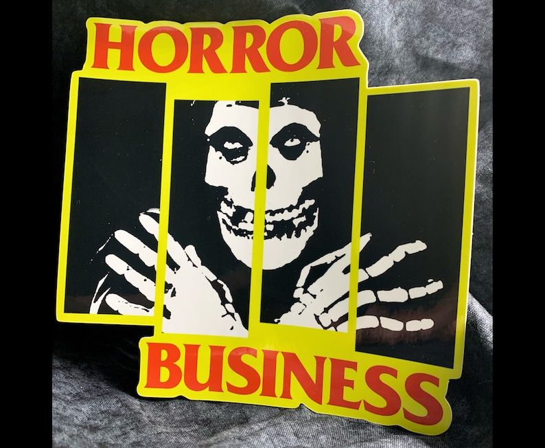 May include: A yellow and black sticker with the words "HORROR BUSINESS" in red letters. A black and white image of a skull with a skeleton hand on either side is in the center of the sticker.