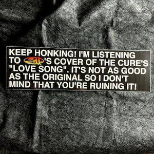 May include: Black bumper sticker with white text that reads "KEEP HONKING! I'M LISTENING TO 311'S COVER OF THE CURE'S 'LOVE SONG'. IT'S NOT AS GOOD AS THE ORIGINAL SO I DON'T MIND THAT YOU'RE RUINING IT!"