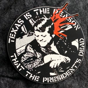 May include: Black and white sticker with a graphic of a man with a red splatter on his head. The text around the graphic reads "Texas is the reason that the president's dead."