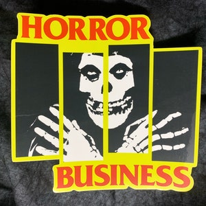 May include: A yellow and red sticker with the words "HORROR BUSINESS" and a black and white image of a skull with a person's hands reaching out from behind bars.
