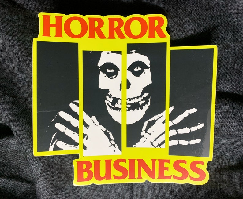 May include: A yellow and red sticker with the words "HORROR BUSINESS" and a black and white image of a skull with a person's hands reaching out from behind bars.