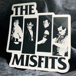May include: Black and white sticker featuring the band The Misfits. The sticker has a black and white image of the band members with the band name in white letters.