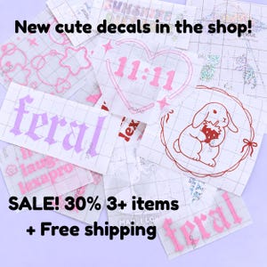 May include: A collection of various vinyl decals in pink and purple. Designs include the word "feral", a heart with the numbers "11:11", and a bunny holding a strawberry. The text "New cute decals in the shop!" is at the top.