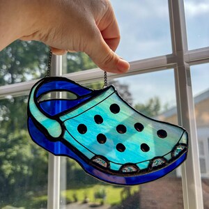 May include: A stained glass suncatcher shaped like a clog shoe. The piece features a light blue iridescent body with dark blue trim and a pivoting heel strap. It includes black circular cutouts and small decorative textured glass accents along the sole.