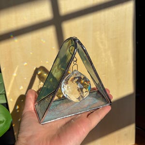 May include: A triangular stained glass suncatcher featuring iridescent green and blue glass panels framed in dark metal, with a clear, multi-faceted crystal sphere suspended from the apex by a small metal ring, catching light to create reflections.