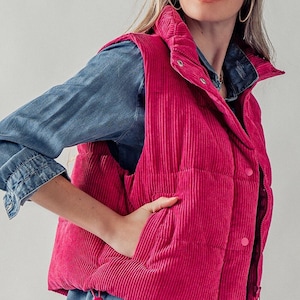 May include: A pink corduroy puffer vest with a zipper closure and button accents. The vest has a stand-up collar and a quilted design.