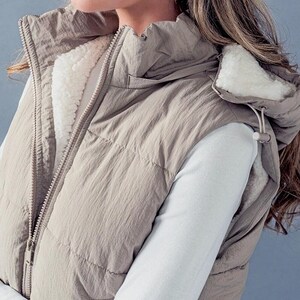 K&ouml;nnte beinhalten: Eine beige gesteppte Weste mit einer Kapuze, die mit Sherpa gef&uuml;ttert ist. Die Weste hat einen Rei&szlig;verschluss und eine Kapuze mit Kordelzug.