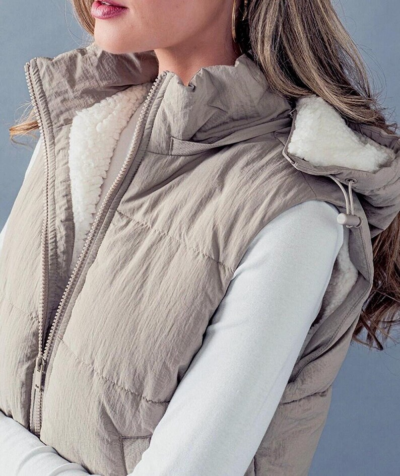 K&ouml;nnte beinhalten: Eine beige gesteppte Weste mit einer Kapuze, die mit Sherpa gef&uuml;ttert ist. Die Weste hat einen Rei&szlig;verschluss und eine Kapuze mit Kordelzug.