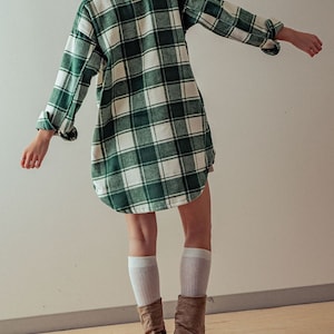 Puede incluir: Una camisa de franela a cuadros verde y blanca con mangas largas y cuello. La camisa se lleva sobre una boina de punto blanca y la modelo lleva botas de ante marr&oacute;n.