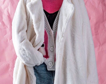May include: A white faux fur coat with a long, loose fit. The coat has a collar and a button closure. The coat is being worn over a pink and gray sweater and blue jeans.