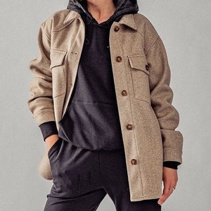 May include: A beige, long-sleeved, button-up jacket with large pockets and a black hooded sweatshirt underneath.