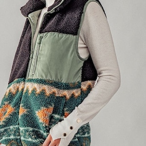 May include: A black, green, and brown patterned fleece vest with a zipper closure. The vest has a high collar and is made of a soft, fuzzy material.