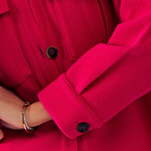 May include: A close-up of a bright pink, long-sleeved shirt with a button-up front and a flap pocket. The shirt has a classic, casual style.