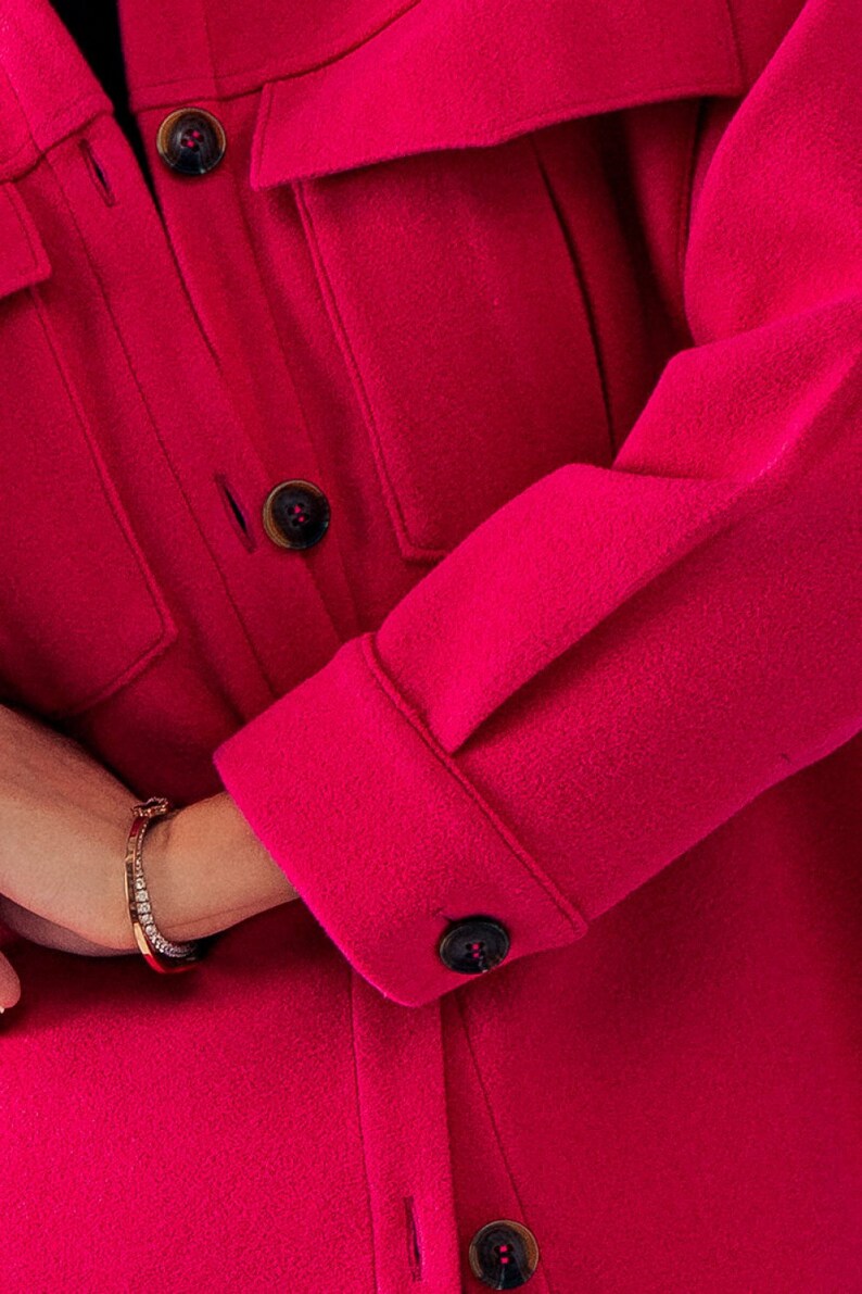 May include: A close-up of a bright pink, long-sleeved shirt with a button-up front and a flap pocket. The shirt has a classic, casual style.