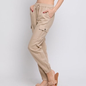 May include: Beige linen cargo pants with a drawstring waist and cuffed legs. The pants have multiple pockets and a relaxed fit.