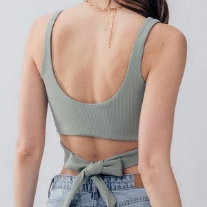 May include: A sage green ribbed crop top with a wrap tie detail in the back. The top is worn with light blue denim jeans.