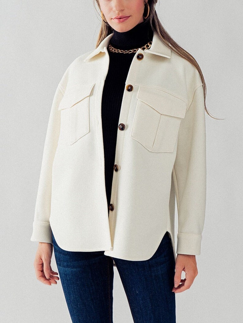 May include: A white oversized shirt jacket with long sleeves, a button-up front, and two large flap pockets. The jacket has a curved hemline and is made of a soft, textured fabric.
