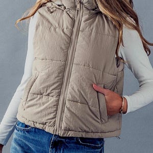 K&ouml;nnte beinhalten: Eine beige gesteppte Weste mit Rei&szlig;verschluss und einer Kapuze mit Kunstfellfutter. Die Weste hat eine lockere Passform und ist perfekt zum Schichten.