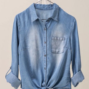 May include: A light blue denim shirt with long sleeves and a button-up front. The shirt is tied at the waist and has a pocket on the left chest.