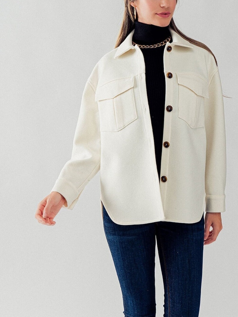 May include: An oversized white button-up shirt jacket with two large pockets on the front. The jacket has a curved hem and is made of a soft, textured fabric.