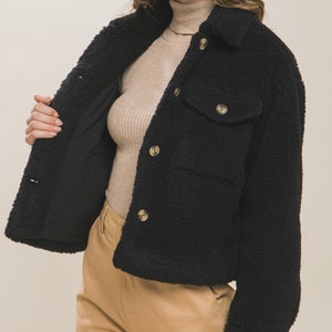 May include: A black cropped sherpa jacket with a button closure and a flap pocket. The jacket has a soft, fuzzy texture.