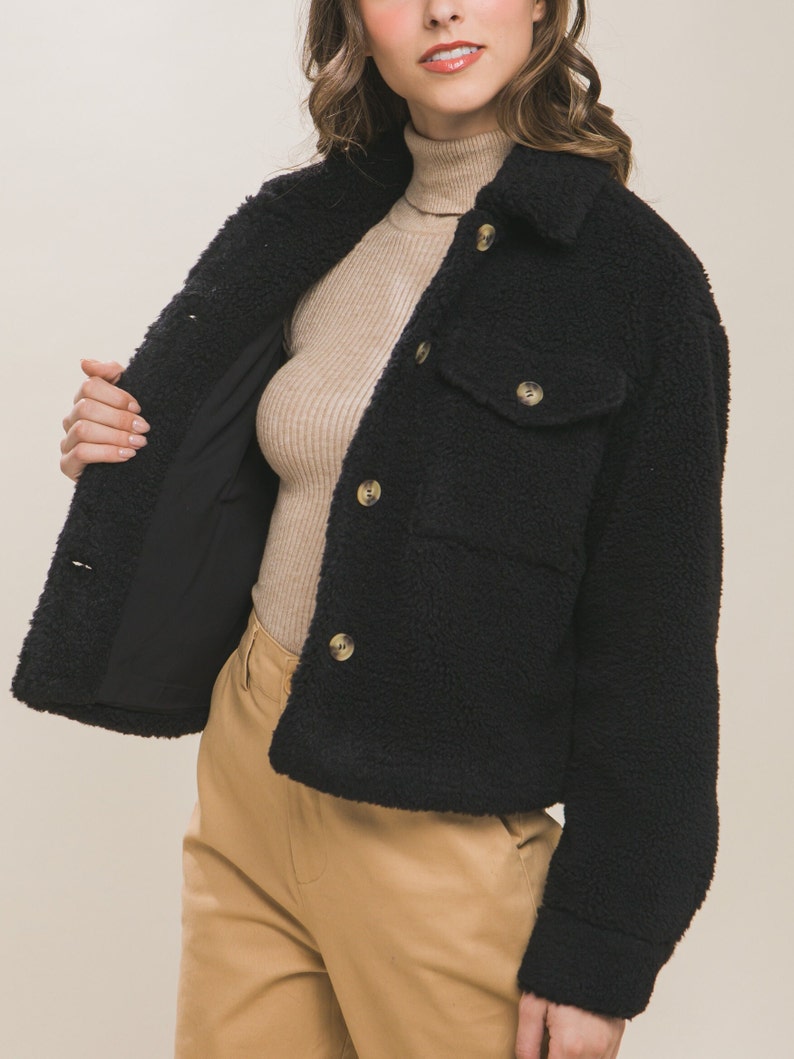 May include: A black cropped sherpa jacket with a button closure and a flap pocket. The jacket has a soft, fuzzy texture.